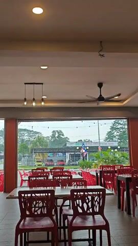 A medium shot captures an empty restaurant interior with red plastic chairs and white tables arranged in rows. The chairs have an intricate back design resembling tree branches. Beyond the restaurant's open front, a street scene unfolds under an overcast sky. A prominent sign with Malaysian flags and text, possibly advertising a business or service, is visible across the street, flanked by trees and parked vehicles. The lighting in the restaurant suggests it is either daytime or evening, with overhead lights and a ceiling fan contributing to the ambiance. The overall impression is one of quiet anticipation, as if the establishment is awaiting customers.