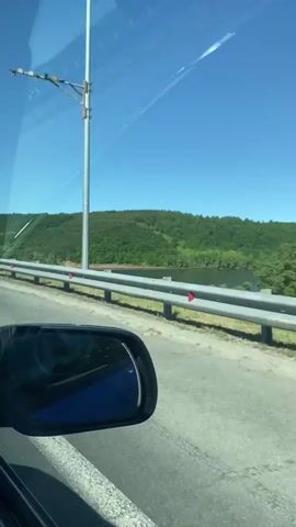 The video shows a car driving over a bridge. A blue sign with white Cyrillic letters reading "Р. ВЯТКА" (R. VYATKA) appears on the right of the road. The river Vyatka is visible to the right behind a guardrail with red and grey supports. Lush green trees fill the far bank of the river, against a clear blue sky. The side mirror of the car is visible in the lower left of the frame.