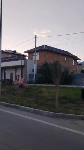 The video captures a view from a moving vehicle, possibly a car, looking towards a street lined with buildings. The sky is overcast with a few wispy clouds, suggesting it's either dawn or dusk. The street is paved, and a curb separates it from a grassy area. In the foreground, a portion of the vehicle's side mirror is visible. Beyond the grass, a low-lying building with a flat roof and several doors is situated. Behind this building, larger structures, including a multi-story brick building, are visible. There's a lamppost in the foreground, and electrical wires crisscross the sky. The camera appears to be panning slightly as the vehicle moves, offering a continuous view of the streetscape. The overall impression is of a quiet, possibly suburban or semi-urban environment during a transitionary period of the day.
