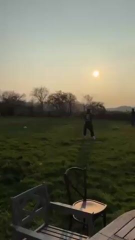 The video shows a grassy field at sunset. In the background, two people are playing with a soccer ball. One person kicks the ball high into the air. In the foreground, there are two chairs and a wooden bench on a patio. The sun is low in the sky, casting a warm glow over the scene.