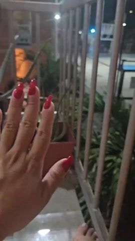 A person stands on a porch at night. The view from the porch through a white metal railing looks out onto an outdoor basketball court illuminated by bright lights. There are potted plants next to the railing. A hand with red, square acrylic nails is brought into view, with the nails clearly displayed. The person wears a beaded bracelet on their wrist.