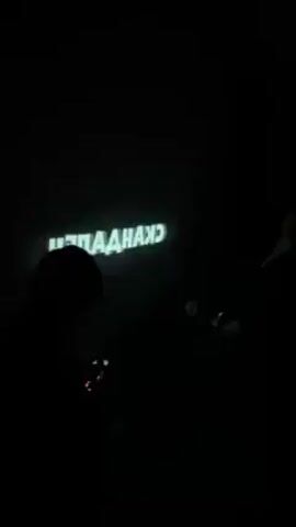 The video shows a person sitting in a chair in a cluttered room with a projector displaying text on a wall. The person is wearing a black hooded sweatshirt and sunglasses. They appear to be watching the projection, and two other people are recording the scene with their phones. The room has boxes and other items scattered around, and the lighting is somewhat dim.