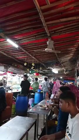 The video depicts the interior of a bustling open-air restaurant or food stall. The ceiling is made of bamboo with red and green tarpaulin stretched across it. Numerous ceiling fans are visible. People are seated at metal tables, eating. Several men are working behind food preparation counters, handling food items and interacting with customers. Blue plastic barrels are scattered throughout the space, seemingly used as support for the food stalls. There are also various containers, buckets, and cooking utensils around the food preparation area. The atmosphere is lively and crowded.