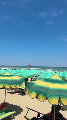 This is a vertical video of a beach scene on a sunny day. In the foreground, there are several brightly colored beach umbrellas, mostly green and yellow, some with a striped pattern. Behind these, the ocean is visible with moderate waves and a clear blue sky. In the mid-ground, a single red umbrella stands out, positioned on a raised platform where a person, likely a lifeguard, is seated. A sign with Italian text is partially visible below the red umbrella. The beach itself is sandy, and some lounge chairs are visible tucked under the umbrellas. The overall impression is a lively and sunny day at the beach.