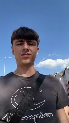 This is a short video captured outdoors on a sunny day in Dobřany, Czechia. The primary subjects are a young man and a young woman walking together. The man is in the foreground, facing the camera, wearing a gray t-shirt with a large white Champion logo. He has dark, bowl-cut hair and is smiling widely at the end of the clip. A black strap from a bag crosses his chest.

The young woman is walking behind and to the right of the man. She has dark hair and is wearing sunglasses and a black top that exposes her left shoulder. She appears to be talking or reacting to something, with her mouth slightly open at one point. She is holding a white bag in her right hand.

The scene is a street with buildings in the background. One building is a light pink apartment building with multiple windows. There are trees and a power pole visible. The sky is blue with scattered white clouds, indicating good weather. The time of day appears to be daytime, likely afternoon, given the bright sunlight and shadows. The overall mood seems lighthearted and casual, with the man's expressions suggesting joy or amusement. The visible text on the man's shirt reads "CHAMPION".