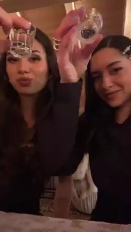 Two young women are in what looks like a cabin with wood beam ceilings. They are holding shot glasses and appear to be making a toast. They then drink their shots, make a face, and look at the camera. They are both wearing dark clothes.