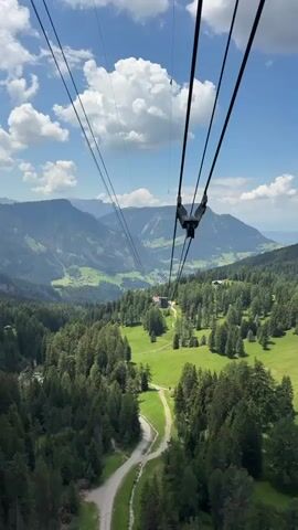 This is a vertical video captured from the perspective of a cable car ascending a mountain. The cable car is the primary object, with its thick black cables dominating the top portion of the frame, stretching diagonally upwards. Suspended from these cables are the mechanisms that hold them in place, and one can see what appears to be a gondola car further up the line, silhouetted against the sky.

The scene below is a breathtaking mountain landscape. Lush green valleys are carved between towering, forested mountains. The dominant colors are vibrant greens from the dense coniferous trees and the rolling meadows, contrasted with the earthy tones of dirt paths winding through the terrain. A gravel road can be seen in the foreground, snaking its way up the mountainside, with a few scattered buildings nestled within the landscape, including a larger structure that appears to be a mountain lodge or restaurant.

The time of day appears to be daytime, indicated by the bright sunlight casting shadows and illuminating the landscape. The weather is fair, with a clear blue sky punctuated by fluffy white cumulus clouds, suggesting a pleasant day for outdoor activities. The location cues of rolling green mountains, dense pine forests, and scattered chalets are consistent with an alpine setting, specifically identified as Urtijëi - St. Ulrich in Gröden - Ortisei, Italy.

The activity taking place is a cable car ride, a mode of transportation commonly used in mountainous regions for both tourism and access. The perspective suggests a journey upwards, offering panoramic views of the surrounding natural beauty. While no people are clearly discernible within the cable car itself, the presence of the cable car implies human activity and tourism. The overall emotion evoked by the imagery is one of awe and wonder at the natural scenery, coupled with a sense of adventure and peaceful ascent. There is no visible text in the media.