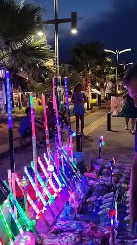 The video shows a night scene on a pedestrian walkway lined with palm trees and streetlights. In the foreground, a stall displays numerous colorful, illuminated novelty items, including what appear to be light-up wands or sticks. Several people are visible in the background, including children and adults. Some individuals are walking along the path, while others appear to be browsing the stall. The atmosphere is lively and festive, suggesting a market or entertainment area.