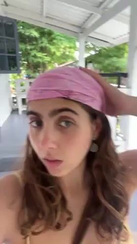 A young woman with a pink bandana and shoulder-length brown hair is standing on a porch, looking directly at the camera. She has fair skin, full eyebrows, and green eyes. She's wearing a yellow tank top and a brown cross-body bag. The porch has white railings and gray painted steps. Trees and other buildings are visible in the background.