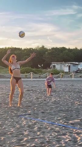 This is a short video of people playing volleyball on a sandy beach. The sun is low in the sky, casting a warm glow. In the foreground, a volleyball net is set up. There are several people on the beach, some of whom are actively playing volleyball. In the background, there are trees and some buildings. The video appears to be taken during the golden hour, with soft lighting and long shadows.