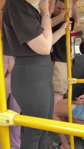A young woman wearing a black cropped t-shirt and black leggings stands on a yellow pole inside a bus. She is holding money in her right hand and looking down at it. Another person wearing a black t-shirt and tan cargo shorts stands behind her. The bus appears to be crowded with other passengers visible in the background.
