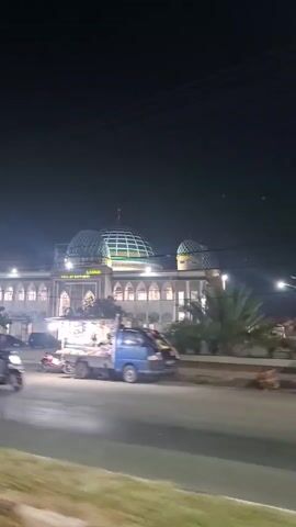 This video shows a nighttime street scene in front of a large, illuminated mosque. The mosque features a prominent green dome and is surrounded by lights. The street is busy with traffic, including several motorbikes, a scooter, a blue van, and at least one car. There are also food carts or stalls set up along the side of the road, illuminated by lights. People are visible riding the motorbikes and scooters, and some are walking near the food stalls. The overall atmosphere appears to be lively and active.