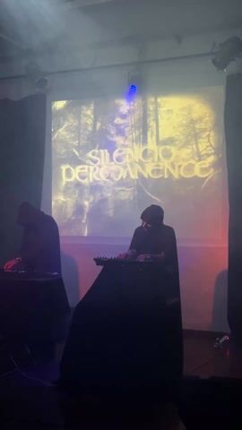 Two hooded figures in black robes stand at tables in a dimly lit space performing music, possibly electronic. They are silhouetted against a projection screen displaying a forest scene and the text “Silencio Permanence” in a stylized font. Spotlights shine down, and there is some smoke or haze in the air. Cables are visible near the tables, and what appear to be stage lights are mounted on the walls.