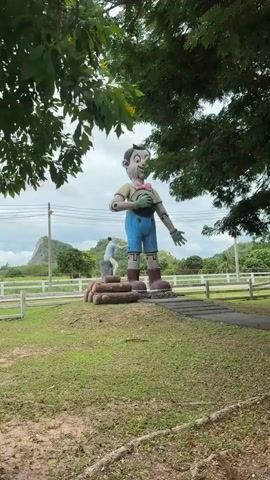 A short video showcases a large outdoor sculpture of Pinocchio, constructed to resemble wood, standing tall against a cloudy sky. Pinocchio is depicted wearing a pink bow tie and blue shorts, with a plaque on his chest that reads "Swiss Sheep Farm". To the left of Pinocchio, a smaller statue of a boy, appearing to be Geppetto, sits on a log pile, reaching up towards Pinocchio's leg. The surrounding environment includes green grass, trees, a white fence, and a distant hillside covered in greenery. The video uses time-lapse or sped-up footage, as indicated by the timestamp.