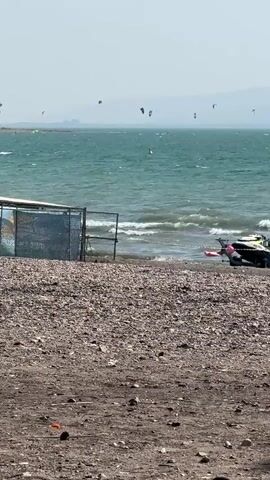 The video shows a beach with choppy, turquoise water. Several kitesurfers are visible in the distance, scattered across the water. On the shore, there is a gravelly area in the foreground, with a structure that appears to be a covered seating area or shelter behind a fence. Part of a boat or personal watercraft is visible on the right side of the frame, near the water's edge. A red inflatable object, possibly a mat or raft, is also in the water close to the shore. The sky is clear and bright, suggesting a sunny day.