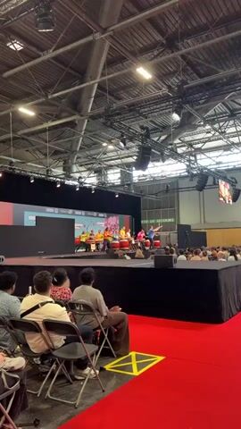 A group of performers, some young and wearing colorful shirts, are on a stage playing large drums. One performer in a blue shirt and black pants is leading the drumming, holding drumsticks aloft. The stage is set against a backdrop with cartoonish imagery and the word "SAKURA" is visible. In front of the stage, an audience is seated in chairs, facing the performance. The venue appears to be a large hall with a high, exposed ceiling and industrial lighting fixtures. A red carpet covers the floor in front of the audience area. Signs indicating an emergency exit are visible in the background.