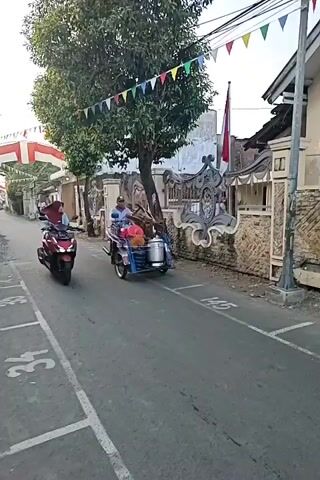A man is riding a modified motorcycle that has a sidecar. The motorcycle and sidecar are blue and the motorcycle is black. The man is wearing a grey and purple shirt and blue shorts. He has a hat on. The sidecar has a large silver container and some red bags in it. The motorcycle is on a paved street in what appears to be a residential area. There are houses and trees in the background. Colorful flags are strung across the street overhead. There are white painted lines on the street, indicating parking spots or lane dividers. A red scooter is parked on the left side of the street, facing the same direction as the motorcycle.