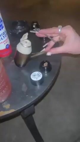 Here is a detailed description of the video:

The video captures a close-up view of a small, round, weathered black metal table with various items related to cannabis consumption. A person’s hands, adorned with elaborate nail art and several rings, are prominent in the frame. The person is using a metal tool resembling tweezers to manipulate a dark brown substance, likely cannabis concentrate, placing it in a small, open container. 

On the table, there is a brown pipe with a white attachment, an open container with dark residue, and a closed container labeled with cannabis warning symbols and text "Strawberry Milkshake".  A can of a pink beverage is also visible. In the background, the environment is dark and appears to be outdoors, possibly on a patio or deck, with indistinct surfaces or structures out of focus.