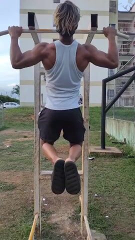 A person with blonde hair is performing pull-ups on a metal bar outdoors. They are wearing a white tank top and black shorts. The background shows a building, grass, and a fence.
