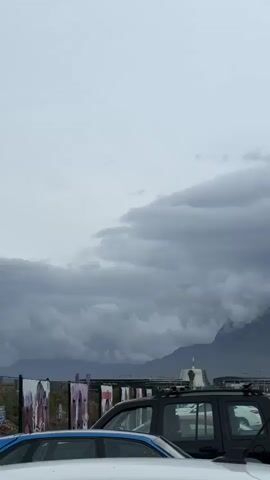 The video shows a cloudy day with a mountain range in the background partially obscured by clouds. In the foreground are parked cars, a bus, and some buildings. The sky is overcast and grey.