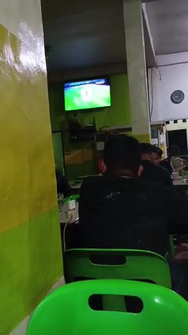 The video shows the interior of a room, likely a cafe or restaurant, with a television mounted on the wall displaying a soccer game. Several people are seated at tables, facing away from the camera, watching the television. The room has green and yellow walls and green plastic chairs. The lighting is dim, with a bright overhead light in the background. The overall atmosphere appears casual and relaxed, with people engaged in watching the game and possibly socializing.