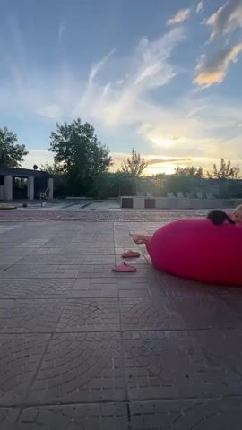 A low-angle shot captures a young girl and a black cat relaxing on a large, bright pink beanbag chair outdoors. The girl is partially submerged in the beanbag, with her feet and legs visible, and her face peeking out from the top. The cat is perched on the beanbag near her head. They are situated on a tiled patio, with a pair of pink flip-flops lying nearby.

The setting appears to be a backyard or outdoor recreational area, possibly near a pool, given the presence of lounge chairs in the background. A blue slide leads down from a structure further back. The scene is bathed in the warm, golden light of sunset, with the sun low on the horizon and casting long shadows. The sky is adorned with dramatic, wispy clouds, suggesting fair weather. The presence of trees and foliage indicates a natural, possibly rural, environment. The overall mood conveyed is one of leisure and contentment, with the girl and cat enjoying a peaceful moment outdoors as day transitions to evening. No visible text is present in the media.