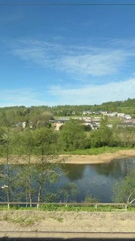 A point-of-view shot from a moving train shows a verdant landscape under a bright blue sky with sparse white clouds. In the foreground, railway tracks with gravel ballast are visible. Beyond the tracks, the scene unfolds with a calm river reflecting the sky, bordered by lush green trees and shrubs. On the opposite bank, houses are nestled on a hillside, surrounded by more greenery. As the camera pans, a wide green field appears, with a path or road winding through it, and a small structure in the distance. The overall impression is one of natural beauty and tranquility in a rural or semi-rural setting. The person filming expresses enjoyment of the view, stating "this is beautiful here."