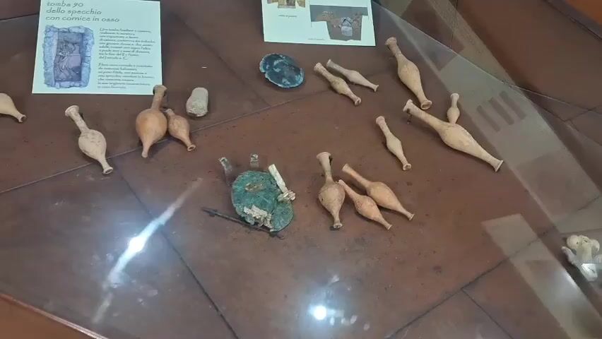 The video shows an archaeological display case with various artifacts. The case has a brown, textured surface, possibly wood, with a glass top. Several small, terracotta-colored vessels of various shapes and sizes are arranged on the surface. Some appear to be amphorae or oil lamps. In the center of the display, there is a greenish, circular object with some remnants of organic material and a metal spike. To the left of this object, there are two more small vessels and a small, gray stone. There is also a flat, dark gray object. Further to the left, a white informational placard is displayed. The placard features an illustration of a sarcophagus or tomb and text in Italian. In the upper right corner of the display, there are additional small terracotta vessels. The lighting in the case is bright, with some reflections visible on the glass surface. The overall impression is that of a museum exhibit showcasing ancient artifacts.