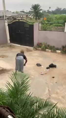 A person is sweeping the courtyard of a building. The person is facing away from the camera, and is wearing a dark shirt and a long, light-colored skirt. They are using a traditional broom made of twigs or branches to sweep litter into a pile. The courtyard is paved with a pattern of square tiles, and there is some trash scattered on it. In the background, there is a dark metal gate, a fence, and some buildings. There are plants along the edge of the courtyard and a palm tree visible. The sky is overcast and appears to be early morning or late afternoon.