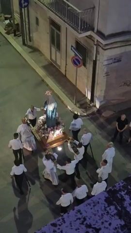A religious procession is taking place outdoors on a street at night. A group of men, most wearing white shirts and dark pants, are carrying a statue of the Virgin Mary on a decorated float. The float is adorned with flowers and has lights. More people, some in white robes, are walking alongside the float and in front of it. Spectators are gathered on the sidewalks and in doorways, watching the procession. Several cars are parked along the street. The buildings on either side of the street are illuminated.