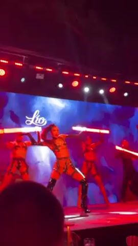 Alright, here's a description of the video:

The video captures a performance on a stage featuring four dancers, who are all dressed in revealing costumes and knee-high boots. They are wielding light sticks, which appear to be red. The stage is brightly lit with a combination of white and red lights. 

Behind the dancers is a large screen displaying a background that appears to be blue with white clouds, and the logo "Lio". The camera angle is from a low vantage point, suggesting the videographer is in the audience. The focus of the video is on the dancers and their performance.