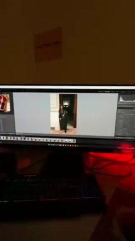 A photograph of a woman wearing a long black robe is displayed on a computer monitor. The woman is standing inside a doorway. A black keyboard is positioned directly below the monitor. A red light is visible behind the monitor. A piece of paper with text in a non-English language is posted on the wall above the monitor. The text appears to read “счастли́вчики.”