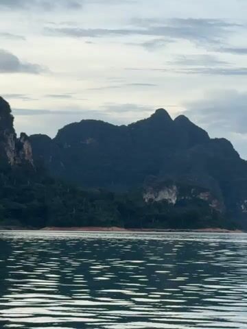 This media file captures a serene boat journey on a calm body of water, likely a lake or reservoir, against a backdrop of dramatic karst mountains. The scene is set at Khao Phang, Thailand, known for its breathtaking natural landscapes.

The primary subjects are the majestic, densely forested karst mountains that dominate the horizon. Their dark, rugged silhouettes are softened by a light cloud cover, suggesting an overcast or early morning/late afternoon sky. The water in the foreground is rippled, reflecting the muted light and creating a dynamic texture that contrasts with the stillness of the mountains. Small, tree-covered islands are visible in the distance, adding to the picturesque scenery.

The activity taking place is a boat ride, implied by the perspective from the water and the movement suggested by the ripples. There are no visible people or objects other than the natural elements. The time of day appears to be during the golden hours, either sunrise or sunset, given the soft, diffused lighting and the dramatic cloud formations. The weather seems calm, with gentle water movement.

Notable details include the dramatic shapes of the karst formations, the lush green vegetation covering them, and the ethereal quality of the light. The overall mood is one of tranquility, awe, and natural beauty. There is no visible text within the media file.