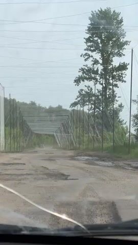 The video shows a car driving along a damaged dirt road, with many potholes filled with water. There are metal poles lining both sides of the road, connected by a grid-like structure above. To the right of the road, there are several wooden Czech hedgehogs (anti-tank obstacles), and a black cross can be seen in the background amongst the trees. The day is overcast, and the windshield wipers are active, suggesting it is raining.