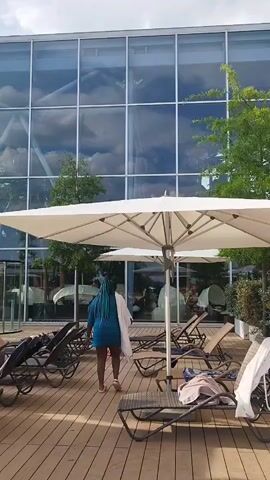 A person with blue hair wearing a teal dress and a white shirt walks away from the camera on a wooden deck. There are lounge chairs and umbrellas on the deck. Behind the deck is a modern building with large glass windows and some trees and bushes. The sky is cloudy.