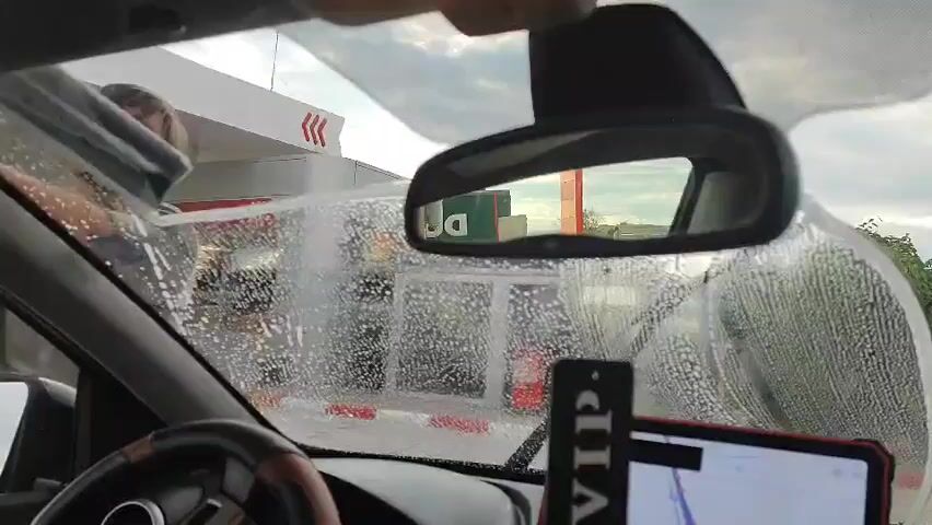 A man with a beard and a grey shirt is standing outside a car and cleaning the windshield with a squeegee. He is at a gas station with a white canopy and red and white striped barriers. The car's interior is visible, including the steering wheel, rearview mirror, and a tablet mounted on the dashboard. The windshield is covered in soap suds. The man is moving the squeegee across the windshield from left to right, attempting to clear the suds. The sky appears cloudy.