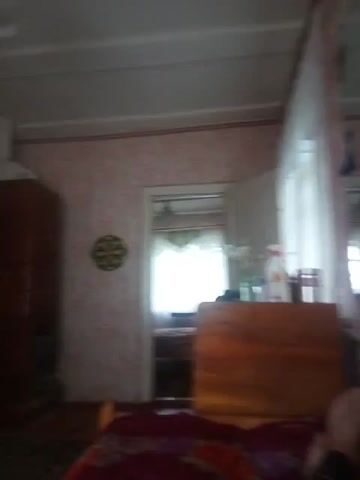 The video shows an indoor scene, possibly a room. The camera is oriented sideways, making it difficult to discern precise details and orientation. A reddish-brown stove or oven is visible in the center of the frame, with a white pipe extending from it. To the left, there's a white vertical object that appears to be a door or panel. The walls are covered with pink and white floral wallpaper. A dartboard is mounted on the wall above the stove. In the background, a mirror reflects a bright light source, likely a window, and hints at furniture. To the right, a corner of a dark-colored piece of furniture and a red patterned blanket or rug are visible. The overall lighting is dim, with a prominent light source coming from the reflected window.