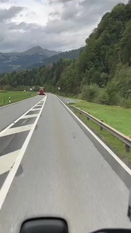 A first-person perspective video shows a road with a red truck in the distance. The road is paved and has white lines indicating lanes. To the right of the road is a metal guardrail and a grassy embankment. Beyond the embankment, there are trees and a lush green hillside. In the far distance, mountains are visible under a cloudy sky. The video appears to be taken from a moving vehicle.