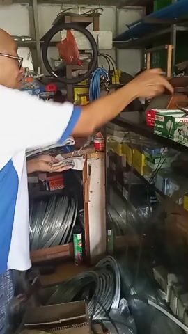 A man wearing a white shirt with blue trim and patterned shorts stands in what appears to be a cluttered workshop or store. He is holding a small object, possibly a piece of metal or wire, in his hands and is examining it closely. The background is filled with shelves stocked with various items, including coils of wire, boxes, tools, and what looks like a bicycle wheel hanging from the ceiling. The lighting is somewhat dim, suggesting an indoor environment. The man's face is partially visible, and he is wearing glasses. He seems to be focused on the item in his hands, possibly inspecting it for quality or damage.
