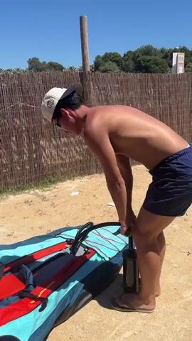 A young man with tan skin, wearing dark blue shorts, light-colored flip flops, a white baseball cap, and sunglasses, is seen from the side pumping air into what appears to be an inflatable kayak laid out on the sand. He is positioned between the kayak and a reed fence. The kayak is mostly turquoise and navy blue with red pads. The ground is sandy. There is vegetation in the background and a clear, bright, blue sky above. There is also a wooden pole in the background.