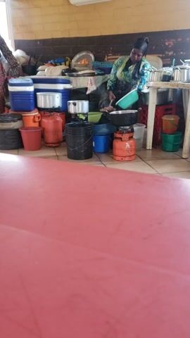 This media file captures a bustling scene in Unguwar Shanu, Nigeria, likely during daylight hours. The primary subject is a woman, dressed in a vibrant, patterned top and a black headscarf, actively engaged in an activity involving various household items. She is positioned near a collection of gas cylinders, pots, buckets, and plastic containers, some of which appear to be for cooking or water storage. Her focused demeanor suggests she is either preparing for something, organizing, or possibly selling these items.

The setting appears to be an outdoor or semi-outdoor area, possibly a market stall or a home's exterior space. Behind the woman, shelves are visible, laden with more metallic pots, cooking utensils, and colorful buckets (including green and orange ones). Crates and what look like Coca-Cola bottles are also present on the shelves, hinting at a commercial aspect to the scene.

A prominent feature to the right of the frame is a large, smooth, reddish-pink surface, possibly a table or a covered structure, which occupies a significant portion of the image and creates a visual division. The ground is tiled, with visible grout lines.

The atmosphere seems active and perhaps a bit cluttered, indicative of a busy day. There are no clear indicators of weather conditions, but the lighting suggests it is daytime. The woman's expression is neutral or focused, not conveying strong emotions. The overall impression is one of daily life and commerce in a Nigerian community. No legible text is visible in the image.