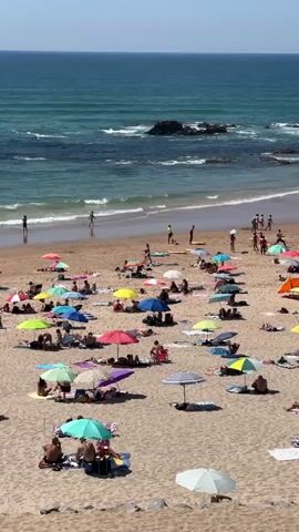 The video shows a crowded beach on a sunny day. The sand is a light brown color, and the ocean in the background is a deep blue with white waves crashing on the shore. Numerous beach umbrellas, in various colors including red, yellow, blue, green, purple, and striped patterns, are scattered across the sand, providing shade for beachgoers. People are seen lounging on towels and blankets, sunbathing, walking, and some are standing or playing near the water's edge. In the distance, there are rocks protruding from the sea. The overall atmosphere is one of a busy summer day at the beach.