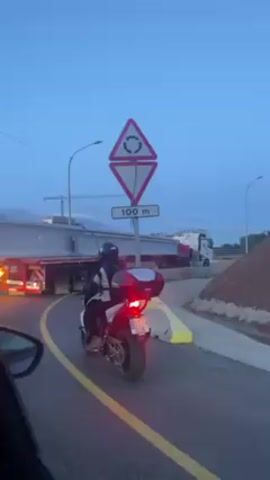A motorcycle is stopped on a curved road, waiting for a wide-load semi-truck to pass. The motorcycle is black and white with a silver rear storage box. The rider is wearing a black helmet and a gray and white jacket. The truck is carrying a large, long, white structure. A traffic sign indicating a roundabout in 100 meters is visible. The sky is blue.