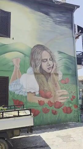 The video pans across a mural painted on the side of a building. The mural depicts a scene with a young girl lying in a field of poppies, with rolling green hills in the background. To the left of the girl, there is a tornado, a picnic basket, and a tin man. Further left, a witch on a broomstick flies across the sky. A small, green, three-wheeled truck is parked in front of the mural. The truck is loaded with green foliage in its bed. The truck also has a painted image of a gnome on its side. In front of the building with the mural, there are two green benches made of wooden pallets, a potted plant with pink flowers, and a metal stand with an umbrella. The ground in front of the building is paved. The video continues to pan to the right, showing more of the building and the surrounding area, including another building with stone walls and a window with green shutters. There is a lamp post and another building in the distance.