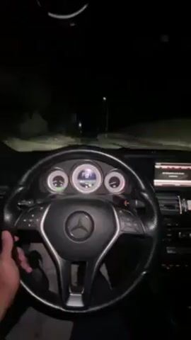 A first-person perspective video shows someone driving a Mercedes-Benz on a dark road at night. The illuminated dashboard and steering wheel are visible, as well as the road ahead illuminated by the car's headlights. Rain or moisture is present on the windshield, and the wipers are in use. Streetlights line the road. The driver's hand is visible on the steering wheel.