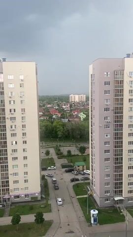 The video shows a view from a high-rise building looking out over a city. In the foreground are two tall apartment buildings, one on the left and one on the right. The building on the left is lighter in color, and the building on the right is a pale purple and cream color. In between the buildings, a street is visible with several cars parked and driving on it. Beyond the street, there are smaller houses with red roofs and green trees. In the distance, the sky is dark and cloudy, suggesting an approaching storm. The overall atmosphere is overcast and somewhat dramatic due to the stormy sky.