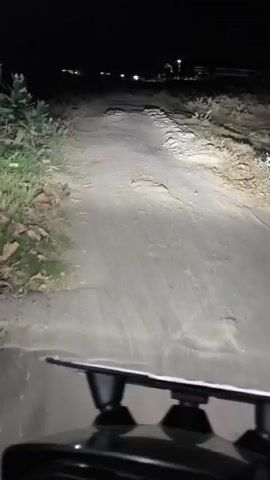 The video shows a first-person perspective of someone riding a motorcycle at night on a dirt road. The headlights illuminate the uneven road surface, which has several potholes and bumps. The rider's digital dashboard is visible, displaying a speed of 14 km/h. The surrounding environment appears to be rural, with sparse vegetation on either side of the road and distant lights in the background. The rider is moving forward, navigating the challenging terrain.