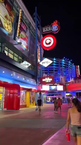 The video shows a night time scene in what appears to be a lively entertainment district, possibly a theme park or a city entertainment area. The camera pans upwards to capture colorful lights illuminating buildings with various restaurants and businesses. There are several people walking in different directions, including a couple holding hands and a woman with a red cup. The video also features escalators leading to higher levels, and signs for Burger Bar, Moe's, Universal Cinema, and food and clubs.
