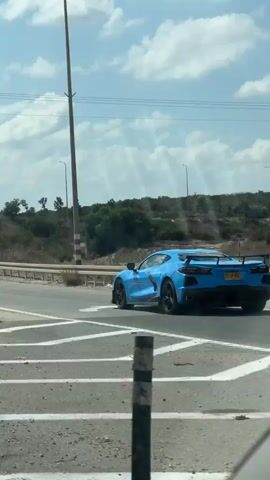 A bright blue sports car is seen driving on a highway under a partly cloudy sky. The car is moving away from the camera and is positioned on the right side of the road. In the background, there are utility poles and signs. One sign has green text in Hebrew and English. Another triangular sign with a red border shows two figures walking. A concrete barrier with metal rails separates the highway lanes from the shoulder, which is covered in dirt and debris.