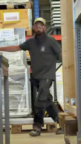 A man with a beard, wearing a dark polo shirt, dark cargo pants, a cap, safety glasses, and dark boots, stands in a warehouse. He has his arms outstretched and his hands open. He moves his right arm and hand towards his face as if to wipe his forehead. He is standing next to a table-like object on his left and a pallet jack on his right. In the background, there are shelves stacked with boxes.